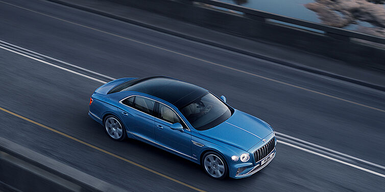 Bentley Al Khobar New Bentley Flying Spur Azure sedan in Light windsor blue paint by Mulliner, with 22 inch Azure wheels seen from overhead driving dynamically on a country road