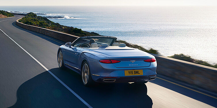 Bentley Al Khobar New Bentley Continental GTC Azure convertible rear view in Light Windsor blue paint with 22 inch ten spoke wheels driving dynamically on an ocean road in the sunshine