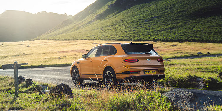 Bentley Al Khobar Bentley Bentayga S SUV rear three quarter in Sunburst Gold paint on a road surrounded by green hills
