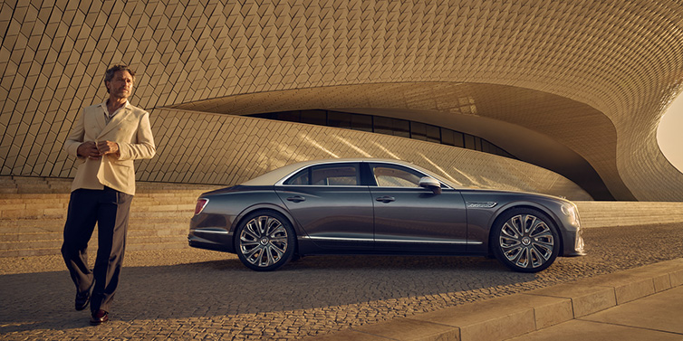 Bentley Al Khobar Bentley Flying Spur Mulliner sedan in Rose Gold over Magnetic duotone paint parked by a curved wall with person in suit jacket standing on the left